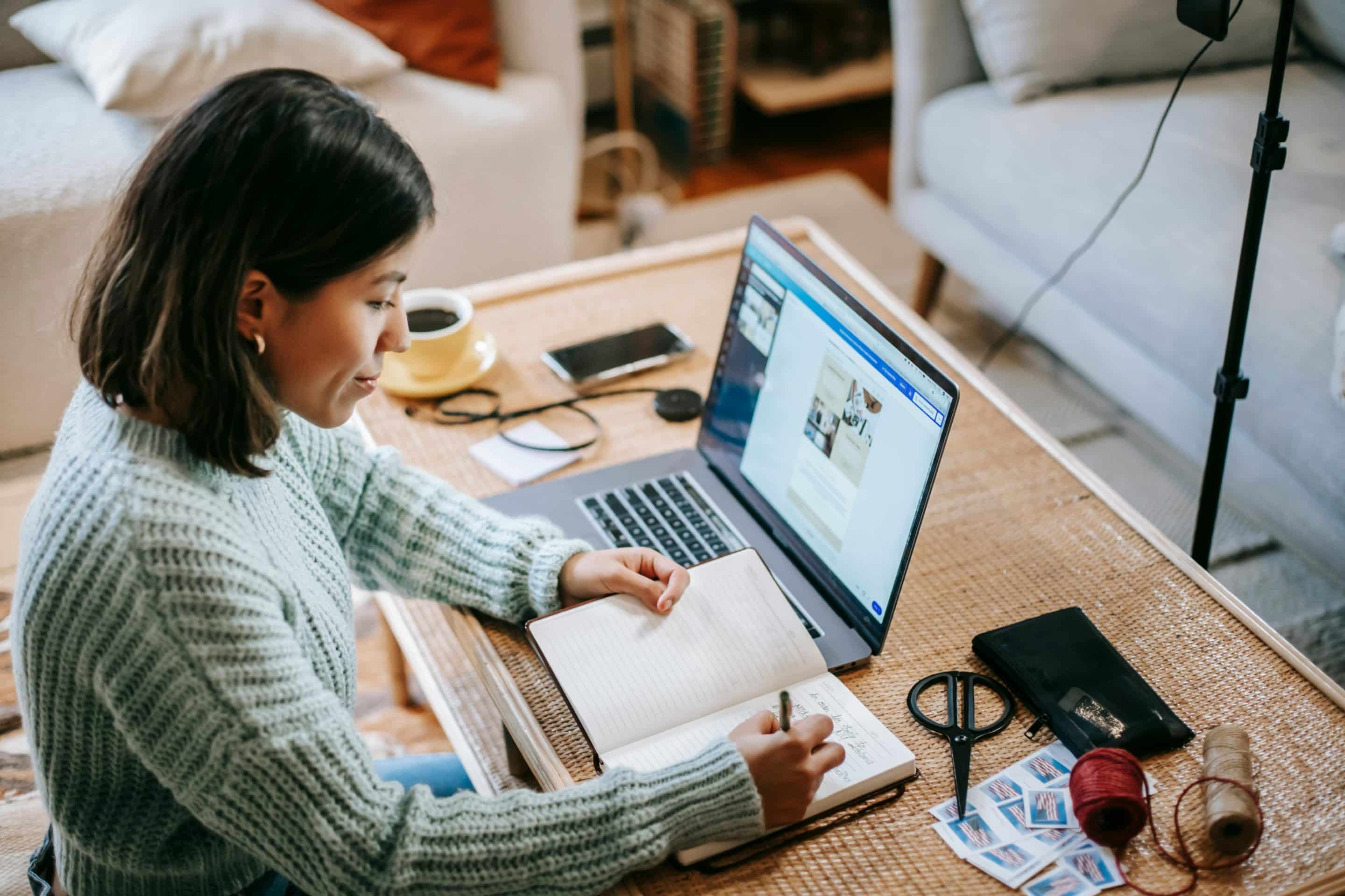 young-focused-woman-writing-in-planner-while-using-laptop